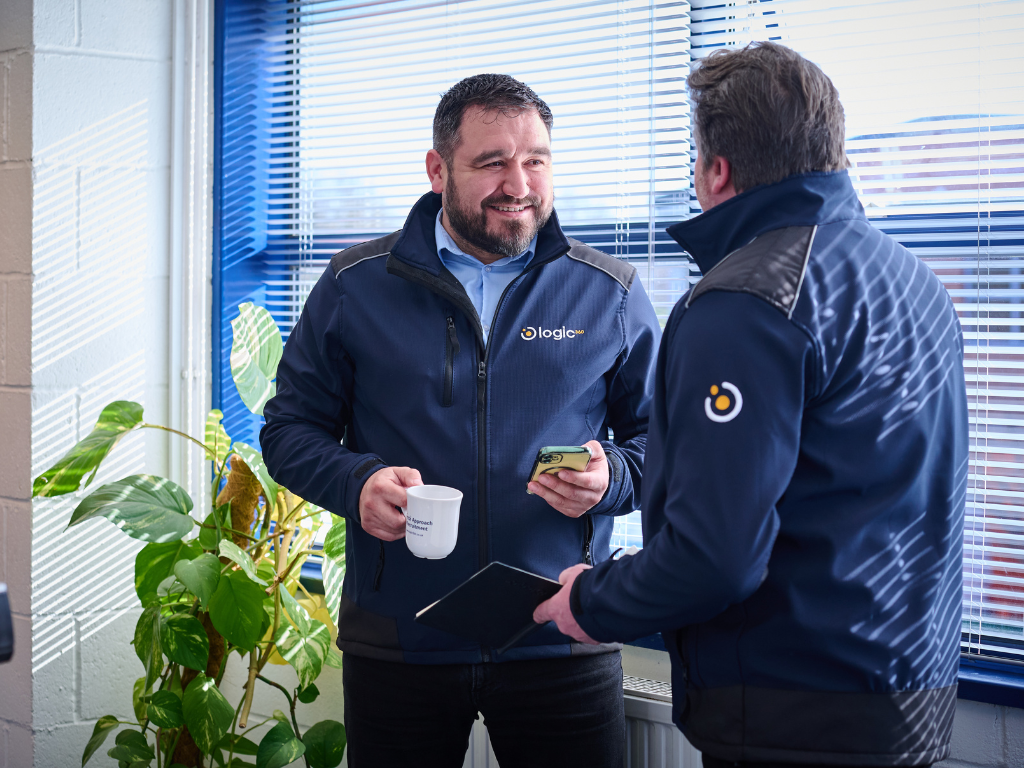 Two Logic 360 team members in branded jackets talking over a coffee in the office.